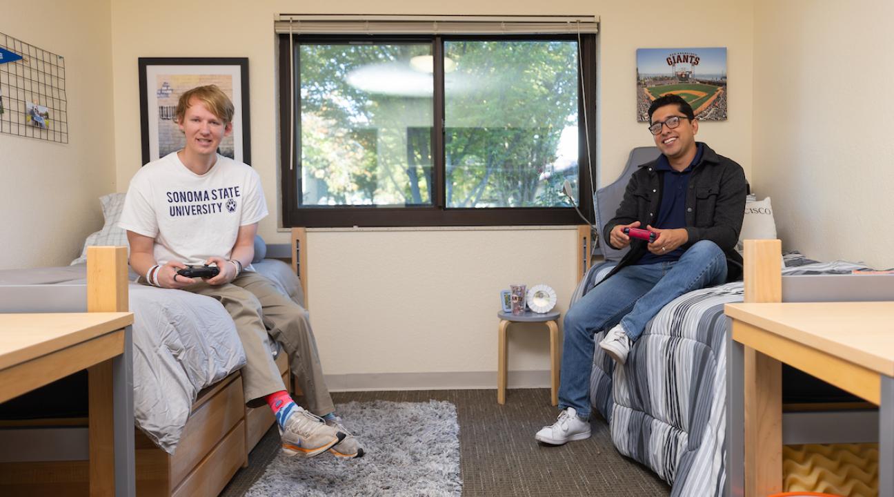 Two students playing video games sitting on their bed