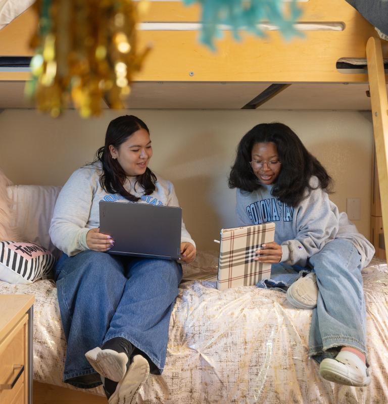 Two students sitting on a bed in a dorm room looking at a computer and folder