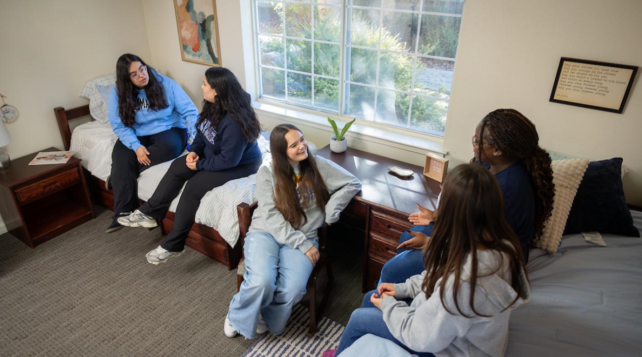 Group of five students sitting on beds in a dorm room socializing