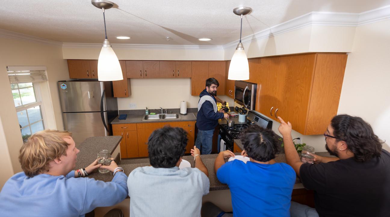 Group of students sitting at kitchen counter while a student cooks at the stove