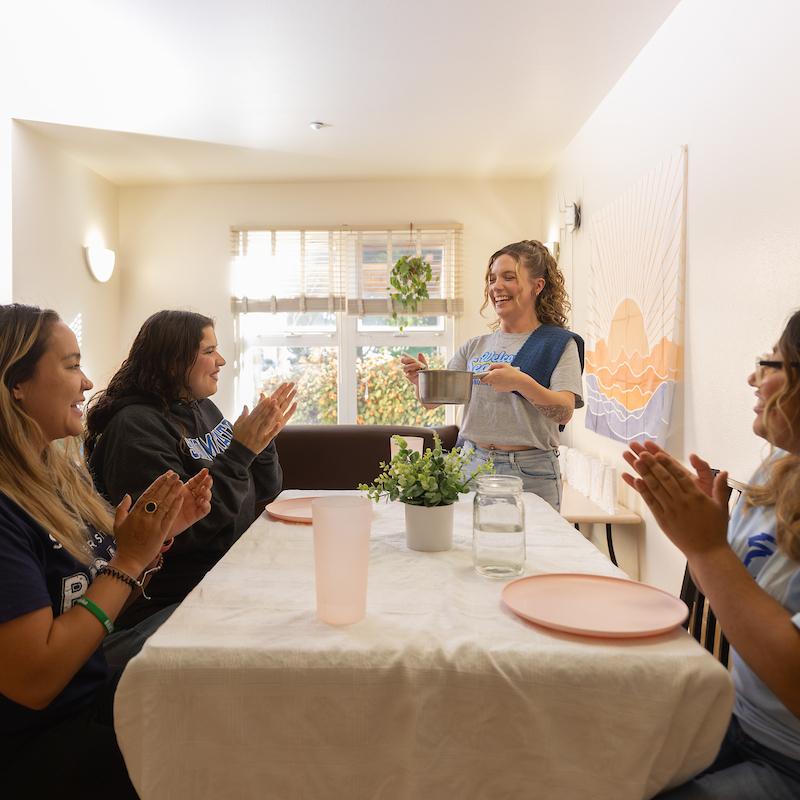 Three students at a dining room tabling with one student serving dinner