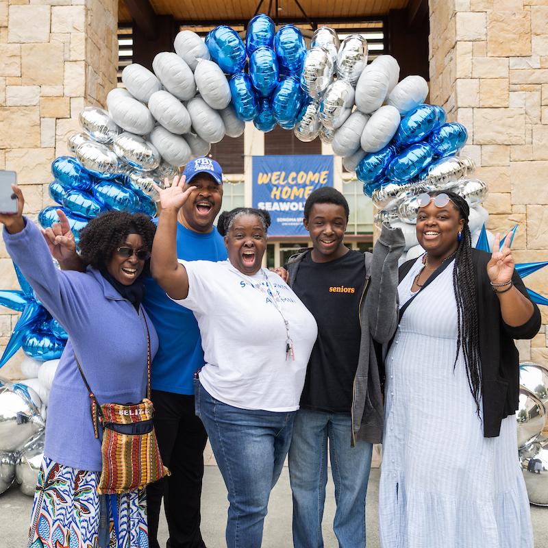 Group of students and parents excited with hands in the air standing in front of a Welcome Home Seawolves banner