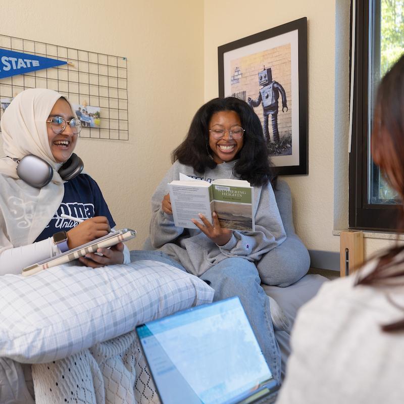 Thwo students sitting on a bed laughing and looking at a book