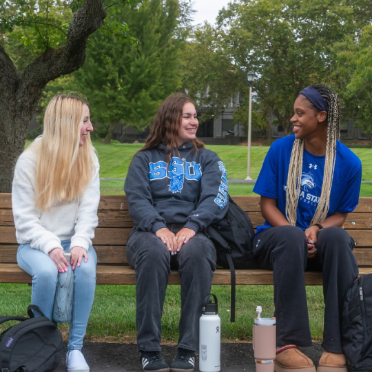 three students sitting on a benchg\