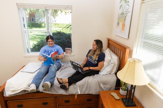 Two students sitting on a bed reading in a dorm room