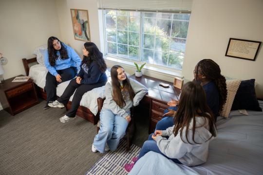 Group of five students sitting on beds in a dorm room socializing