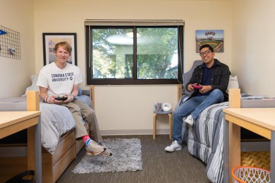 Two students playing video games sitting on their bed