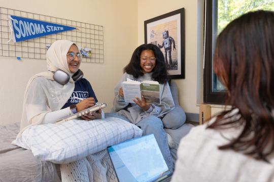 Thwo students sitting on a bed laughing and looking at a book